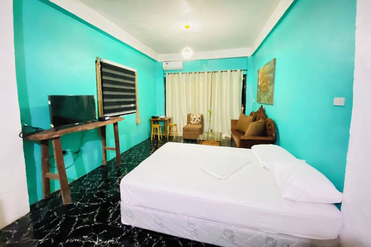 A hotel room with a bed, a desk with a television, a chair, a small table, and a window with curtains.