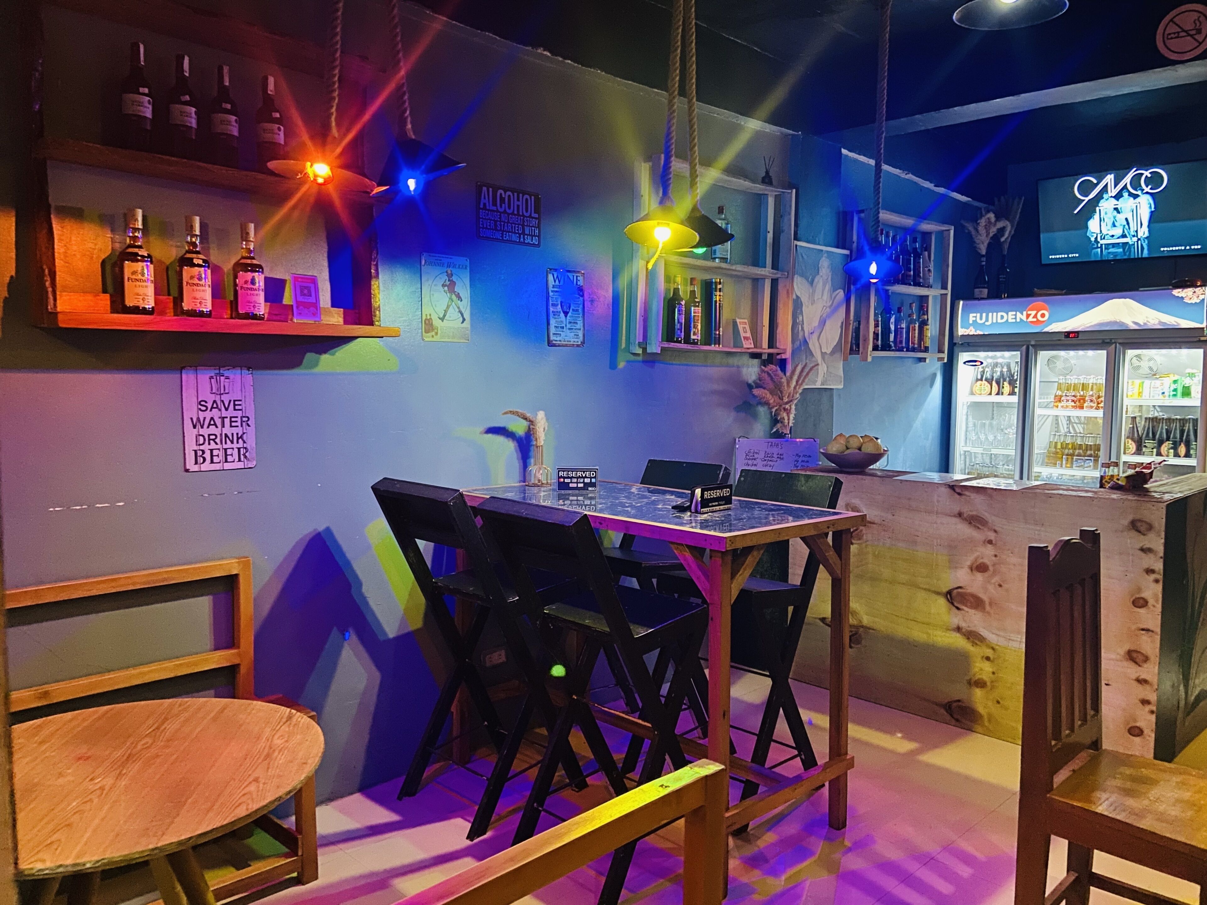 A bar area with a table and chairs, shelves with bottles, and a neon sign.