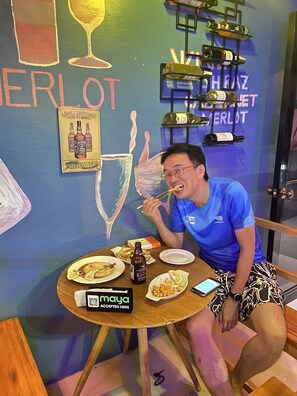 A person seated at a table with plates of food, a bottle of beer, and a smartphone. The wall behind features a mural with wine bottles and the word "MERLOT". - Carib Apartment (Malay)