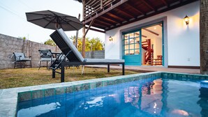 Una piscina con sillas de descanso y una sombrilla, una terraza de madera y un edificio con puerta azul y escalera.