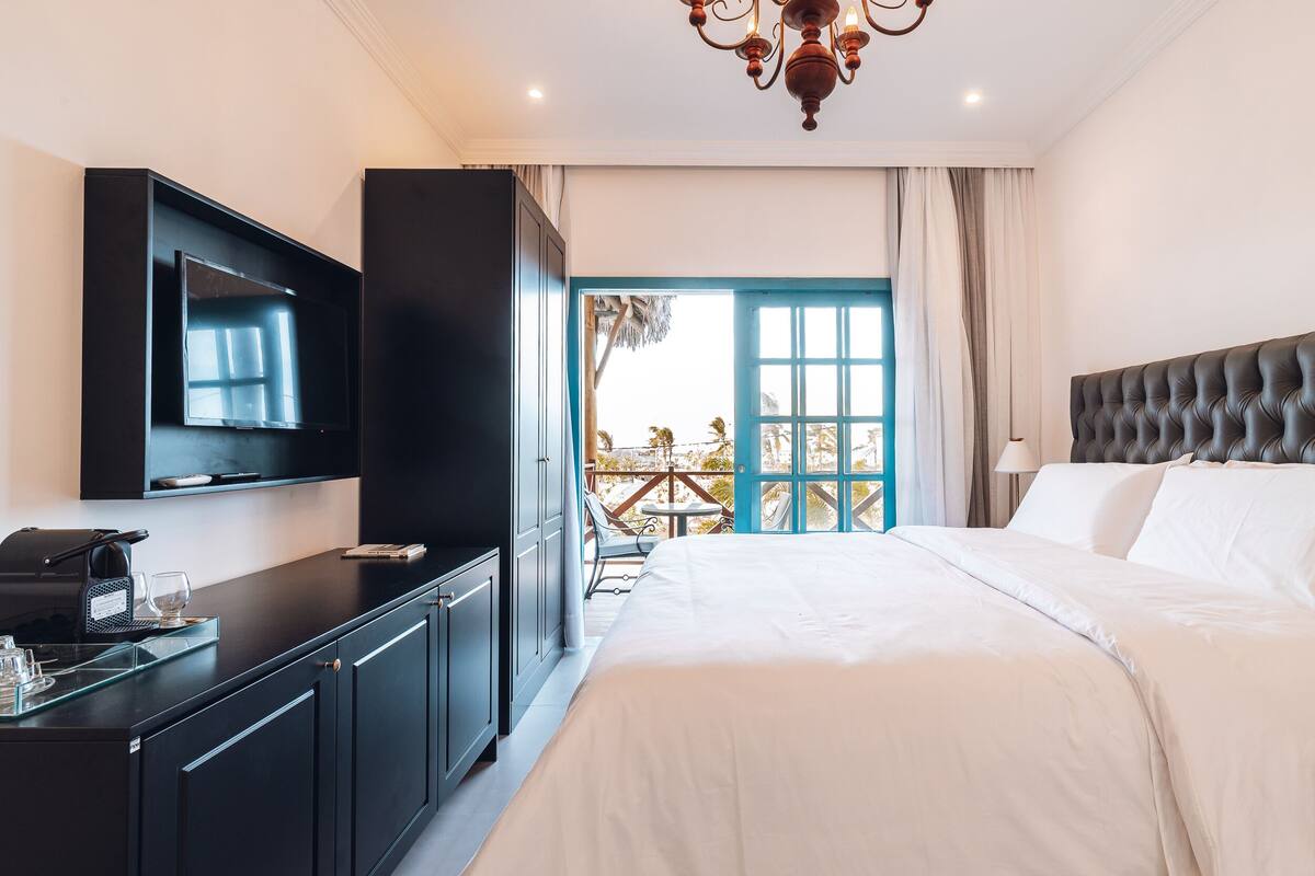 Ein modernes Hotelzimmer mit einem großen Bett, einem flachbildfernseher an der Wand, einem Schreibtisch mit Kaffeemaschine und einem Balkon mit Blick ins Freie.