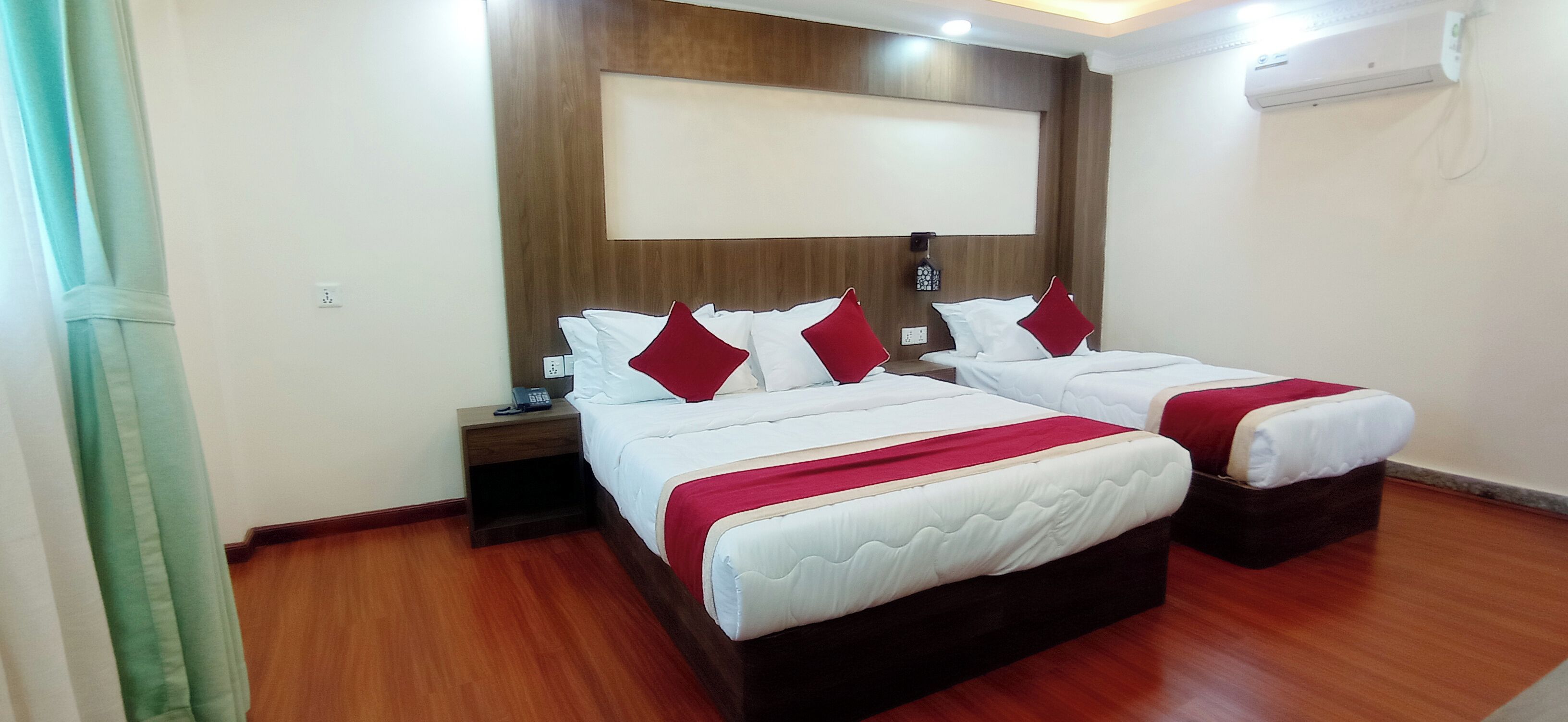 A hotel room with two beds, wooden flooring, a mounted air conditioner, and a wall-mounted light fixture.