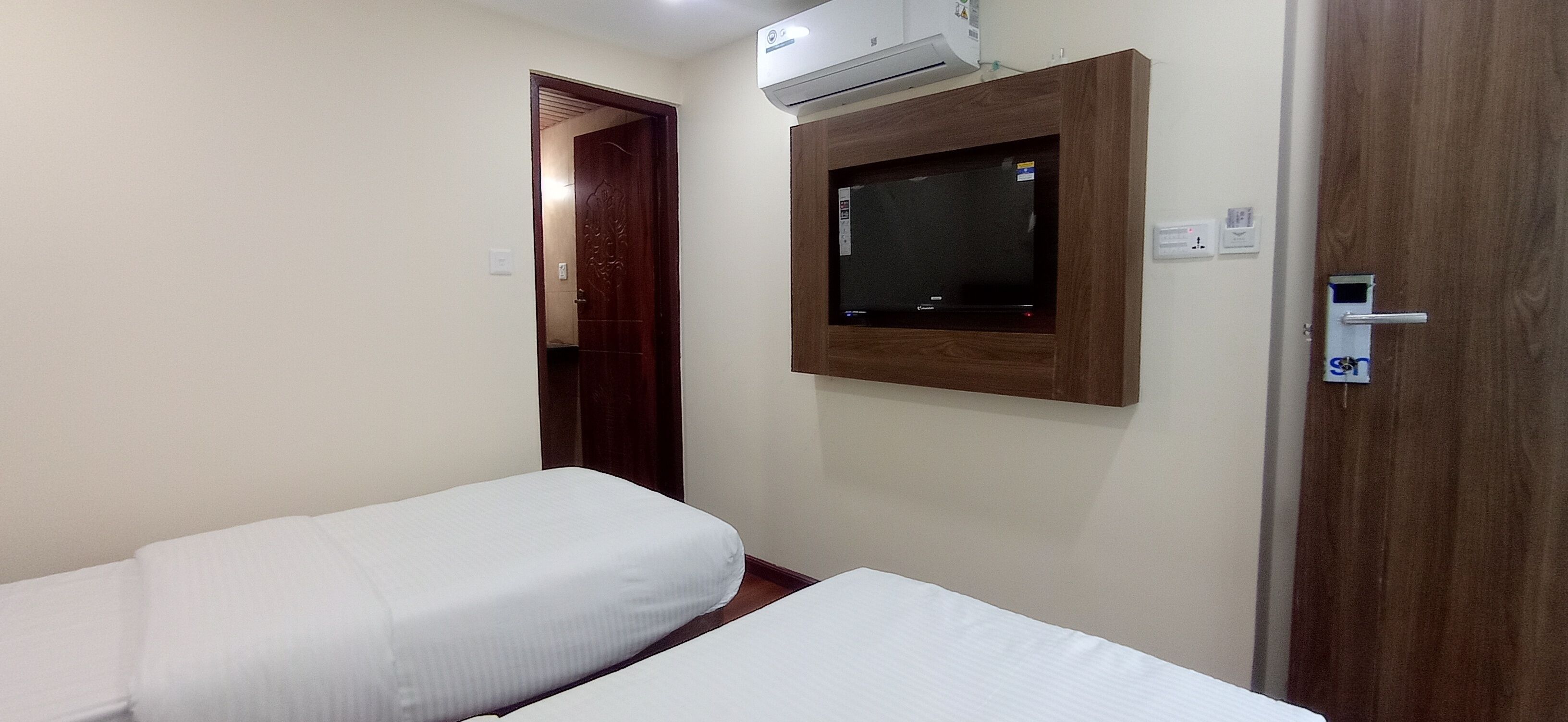 A hotel room with two single beds, a wooden door, a mounted TV, and an air conditioning unit.