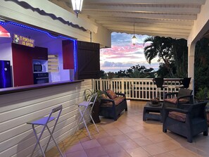 A bar area with a sign indicating it's open, a kitchen with appliances, outdoor seating with chairs and cushions, and a view of the sky and trees.