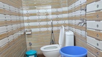 Un baño con piso de madera, un inodoro, un balde azul y un ventilador blanco.