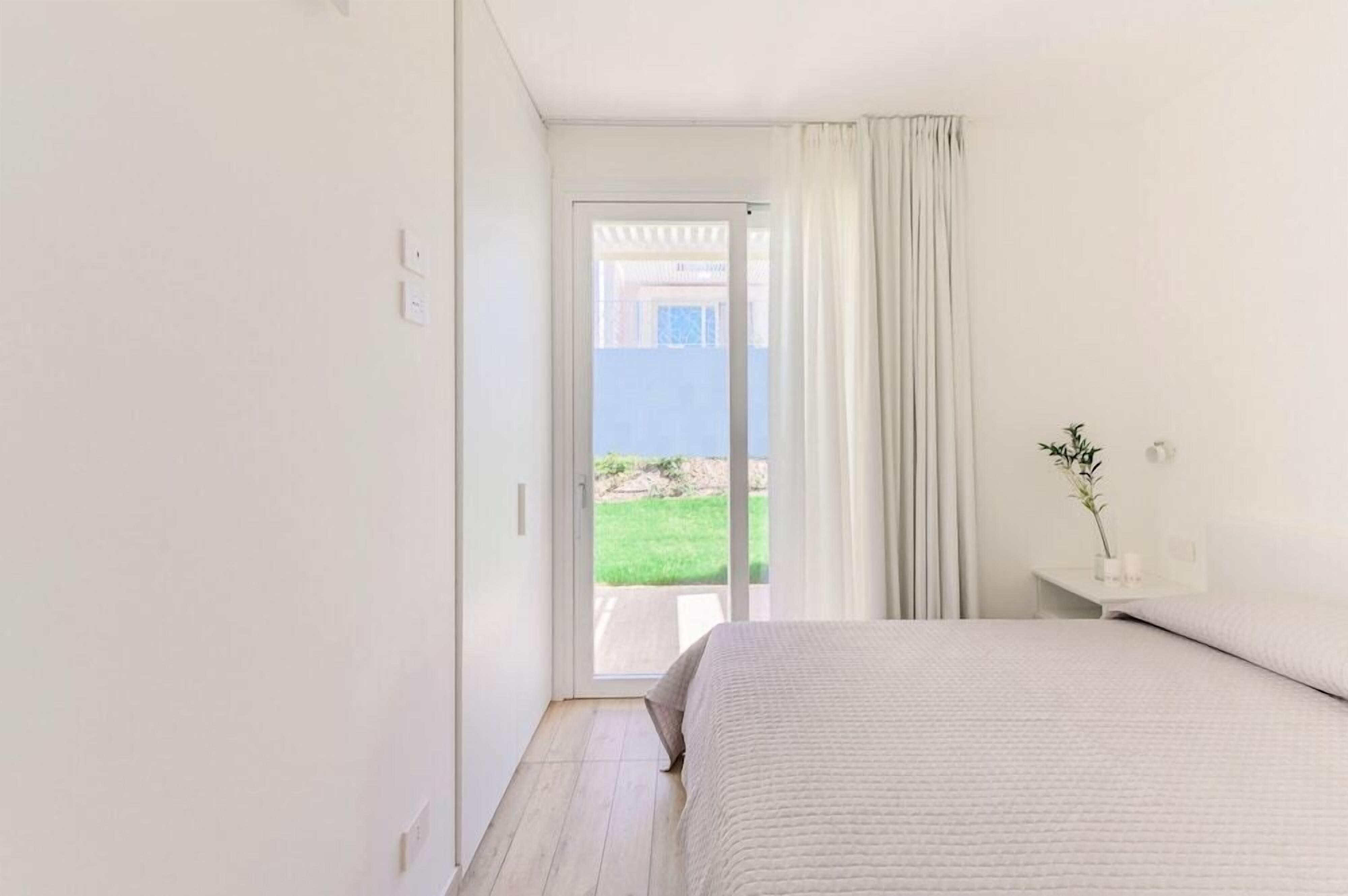 Une chambre à coucher avec un lit, une table de chevet, un vase avec une plante et une vue sur une pelouse verdoyante, visible à travers une porte coulissante.