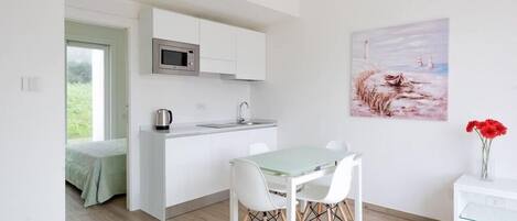 A modern kitchen with white cabinets, a microwave, and a sink. There is a dining area with a table and chairs. A painting of a beach scene is on the wall.
