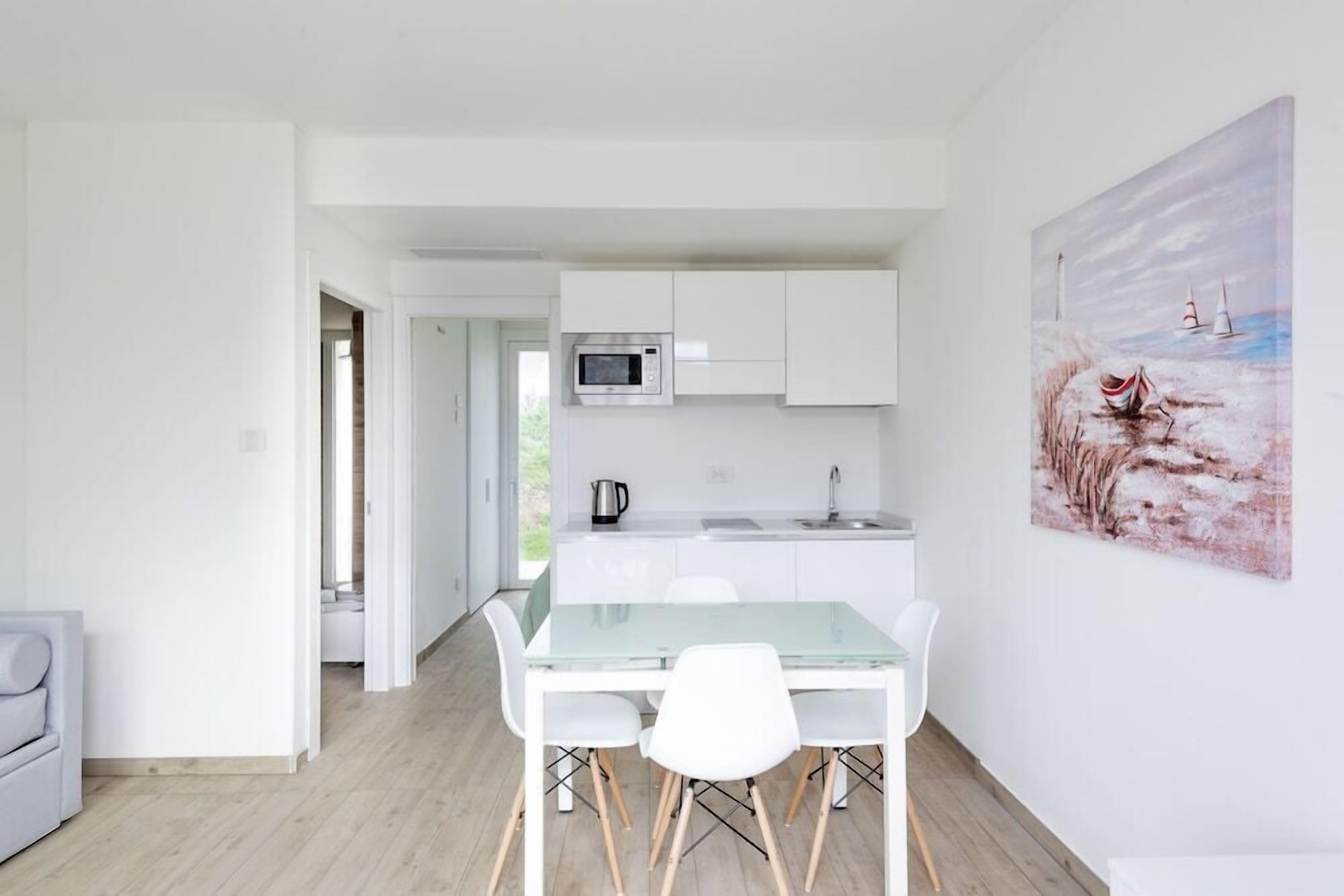 A modern kitchen with white cabinets, a microwave, and a sink. There is a glass dining table with white chairs. A painting of a seascape is on the wall.