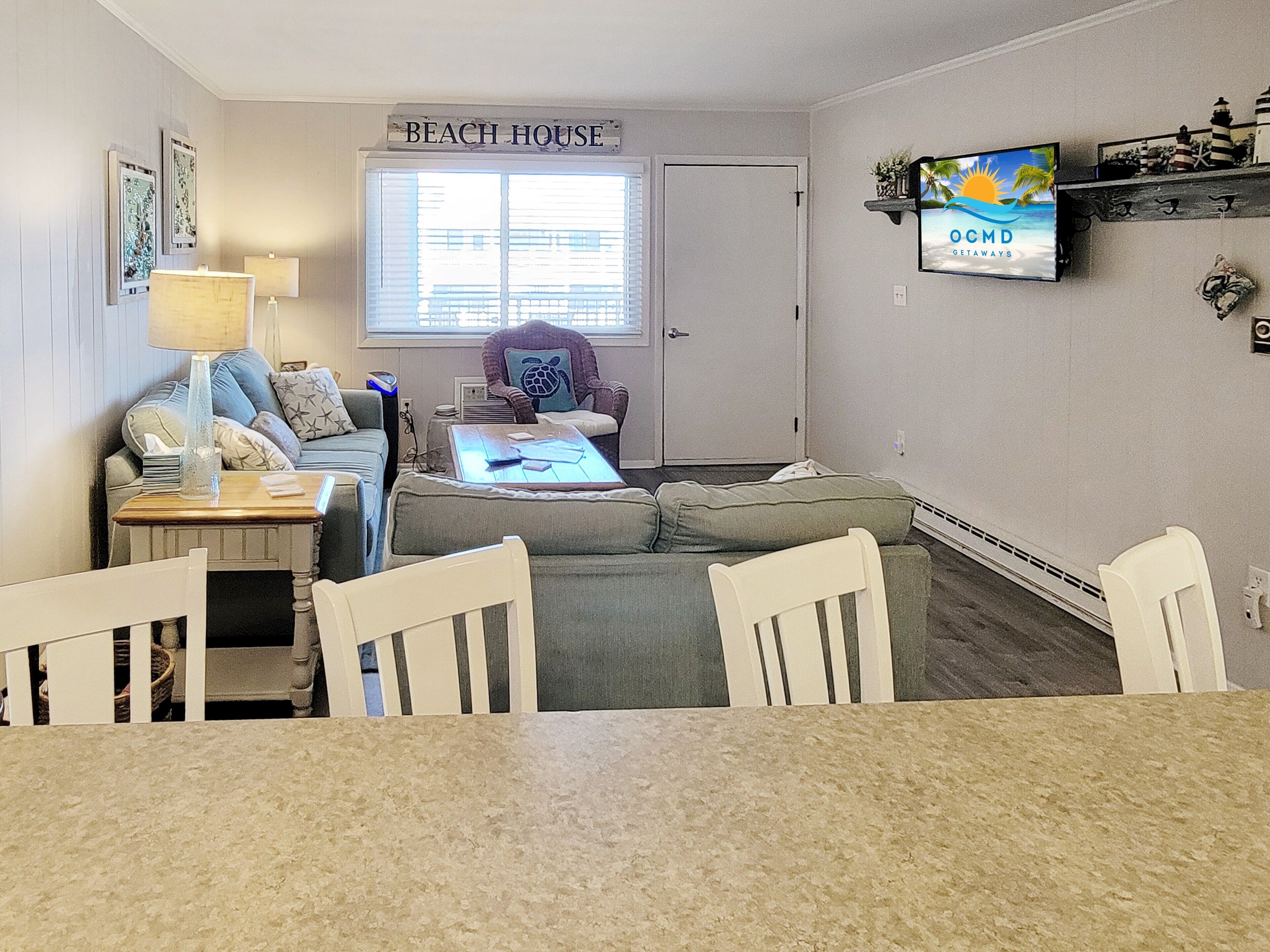 Ein Wohnzimmer mit einer Couch, einem Couchtisch und Sesseln. Ein Schild mit der Aufschrift 'BEACH HOUSE' befindet sich über einem Fenster. Ein Fernseher ist an der Wand montiert.