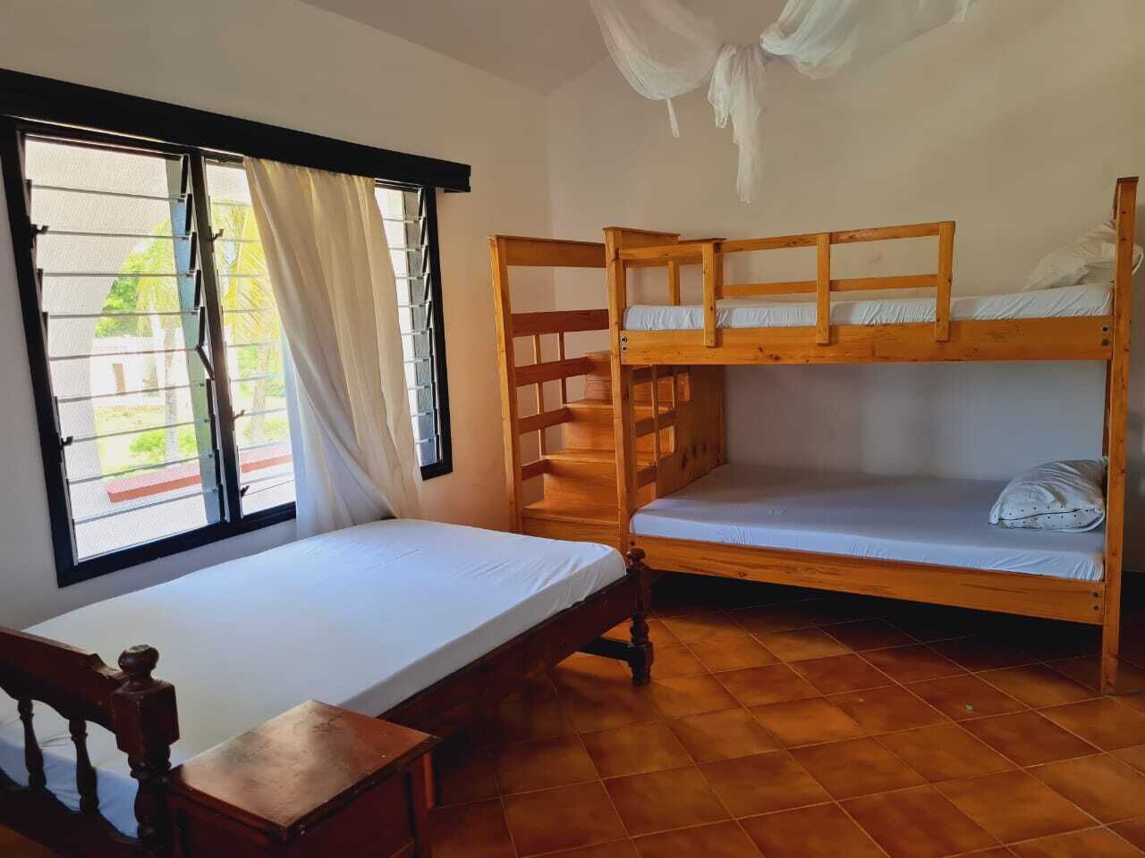 A room with a single bed, a wooden bunk bed, a window with curtains, and a tiled floor.