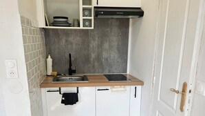 Cozinha compacta com armários brancos, pia de aço inoxidável, coifa preta e tapete cinza estampado na entrada.