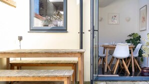 Un banc et une table en bois sont placés à l’extérieur, devant une porte. Une porte vitrée donne accès à une pièce où se trouvent une table et des chaises.