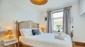 Un lit bien fait, avec du linge de lit blanc et des coussins décoratifs, une lampe de chevet et une petite table sur laquelle se trouve une boîte à motifs à carreaux.