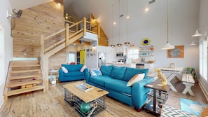 Amplia sala de estar con una escalera de madera, un sofá azul, una mesa de centro de madera y una cocina al fondo.