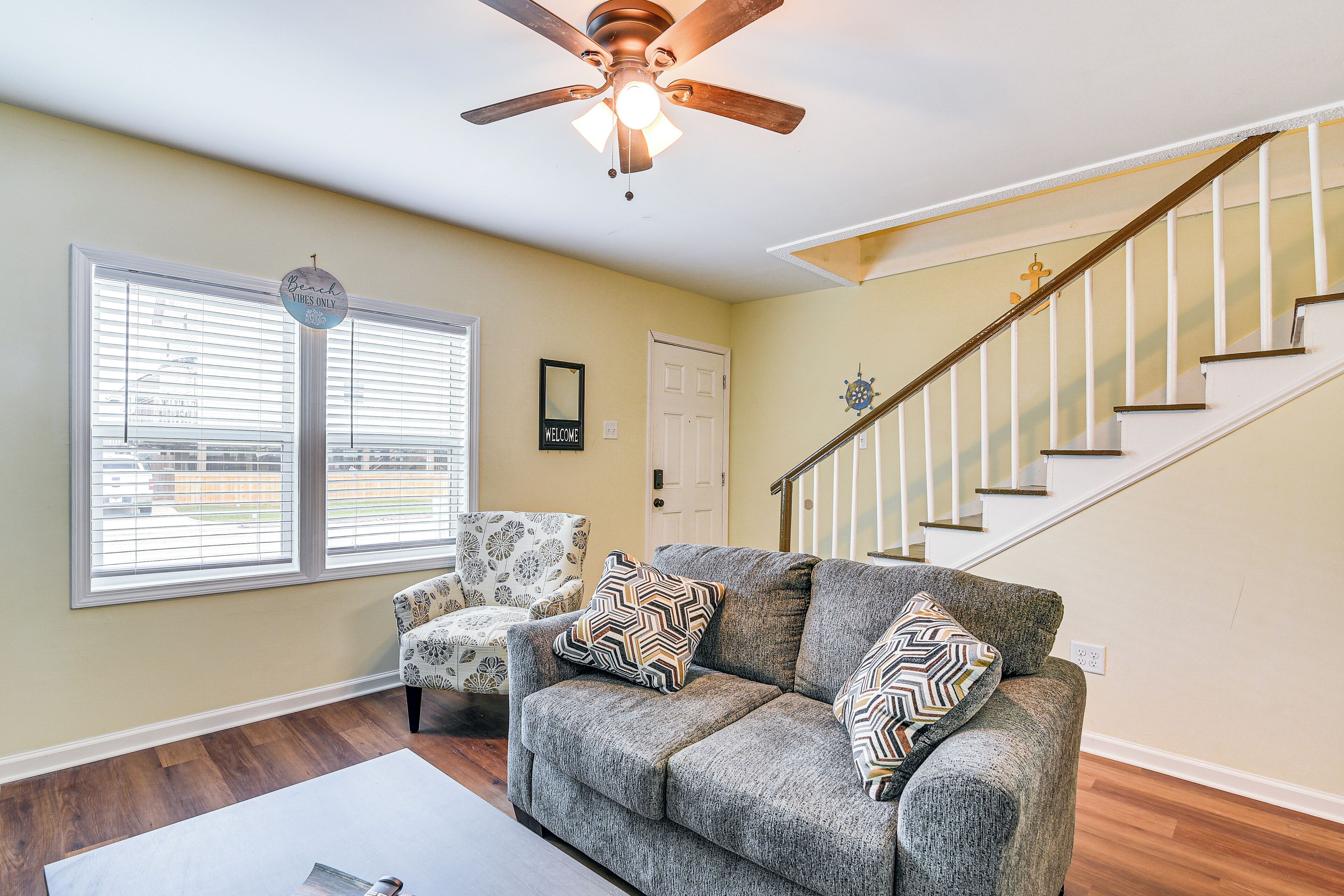 A living room with a grey sofa, patterned pillows, a white armchair, a ceiling fan, and a staircase.
