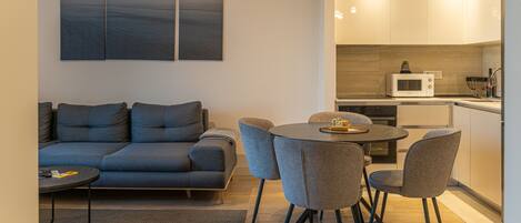 Una sala de estar moderna con un sofá gris, una mesa de comedor redonda con cuatro sillas, y una zona de cocina con microondas.