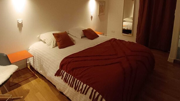 Un dormitorio con techo de madera, cama con ropa blanca y detalles rojos, una mesita con una silla y un espejo en la pared.