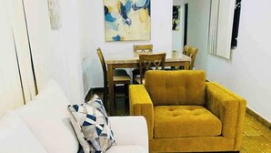 Uma sala de estar com um sofá branco e uma poltrona cor mostarda, uma mesa de jantar com cadeiras e um chão com ladrilhos amarelos.