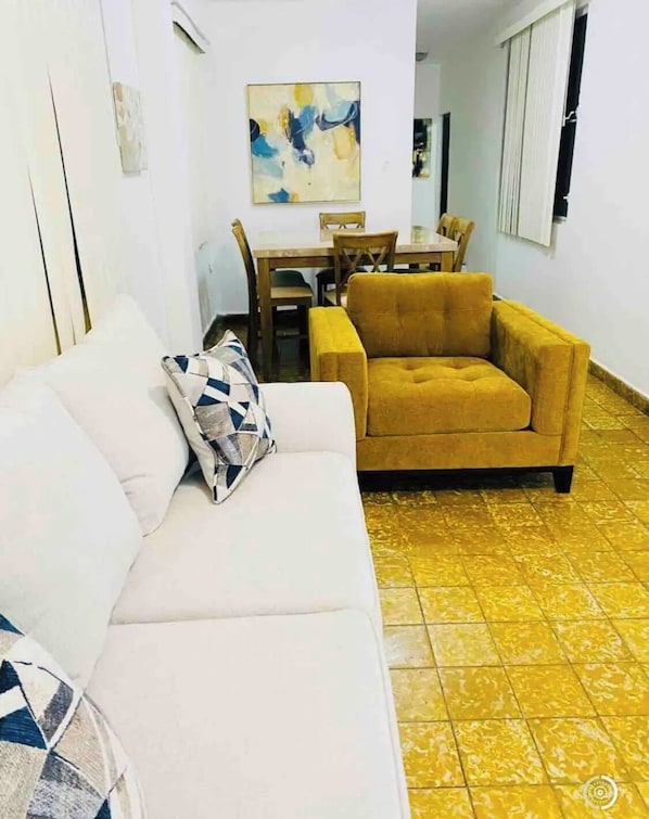 Un salon avec un canapé blanc et un fauteuil de couleur moutarde, une table à manger avec des chaises, et un sol recouvert de carreaux jaunes.