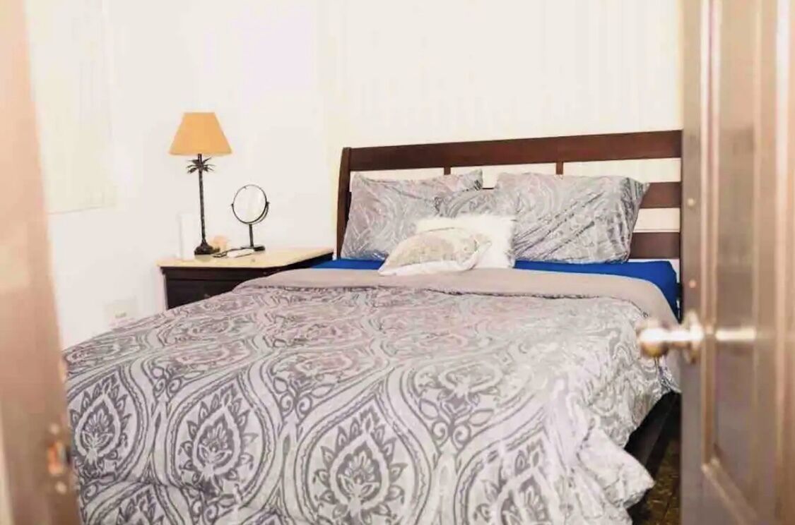 Una cama bien hecha con ropa de cama estampada, una lámpara de noche y una mesita con un espejo.