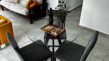 A modern living room with a glass-top coffee table, two grey upholstered chairs, a sofa with patterned cushions, and a potted plant.