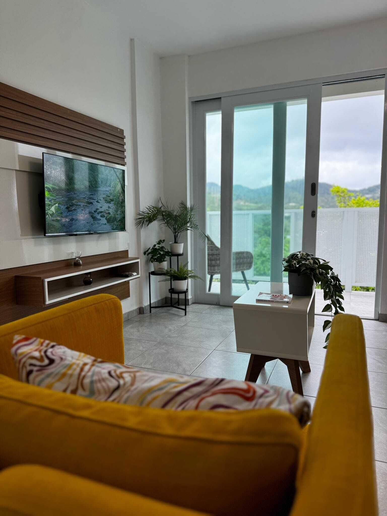 A modern living room with a flat-screen TV, a yellow sofa, a dining table with chairs, and a balcony with a view of greenery and mountains.