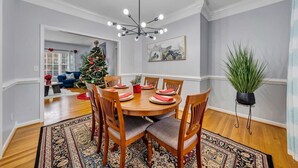 Un comedor con una mesa de madera dispuesta para cuatro personas, rodeada de sillas con asientos acolchados. Un gran macetero se encuentra a un lado.