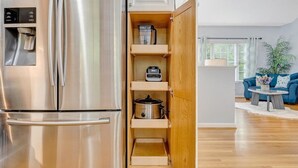 Un refrigerador de acero inoxidable con dispensador de hielo y agua incorporado, un gabinete de madera con estantes y un área de cocina con un sofá azul y una mesa de centro al fondo.