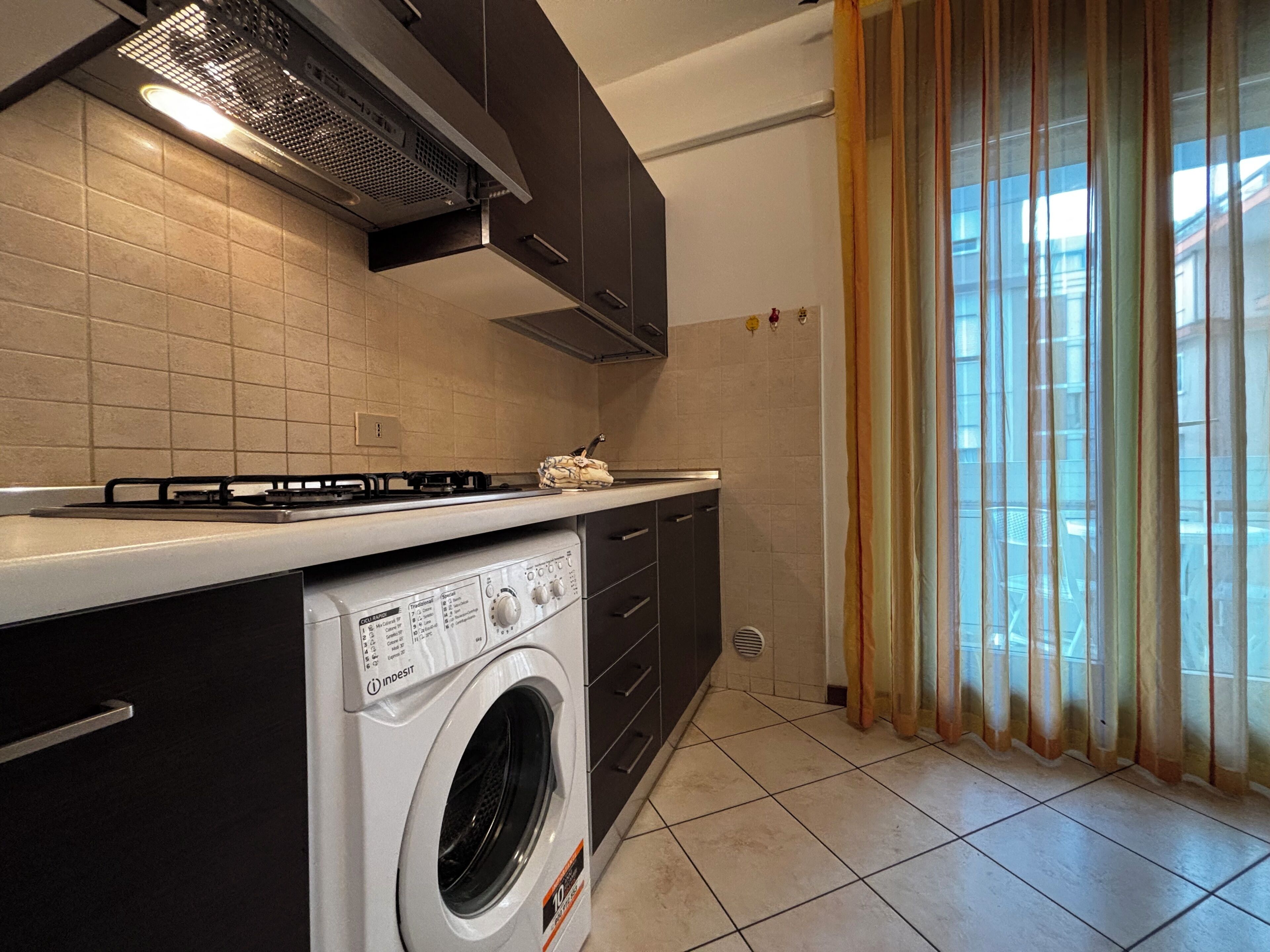 Uma cozinha compacta com fogão a gás, máquina de lavar embutida e janela com cortinas.