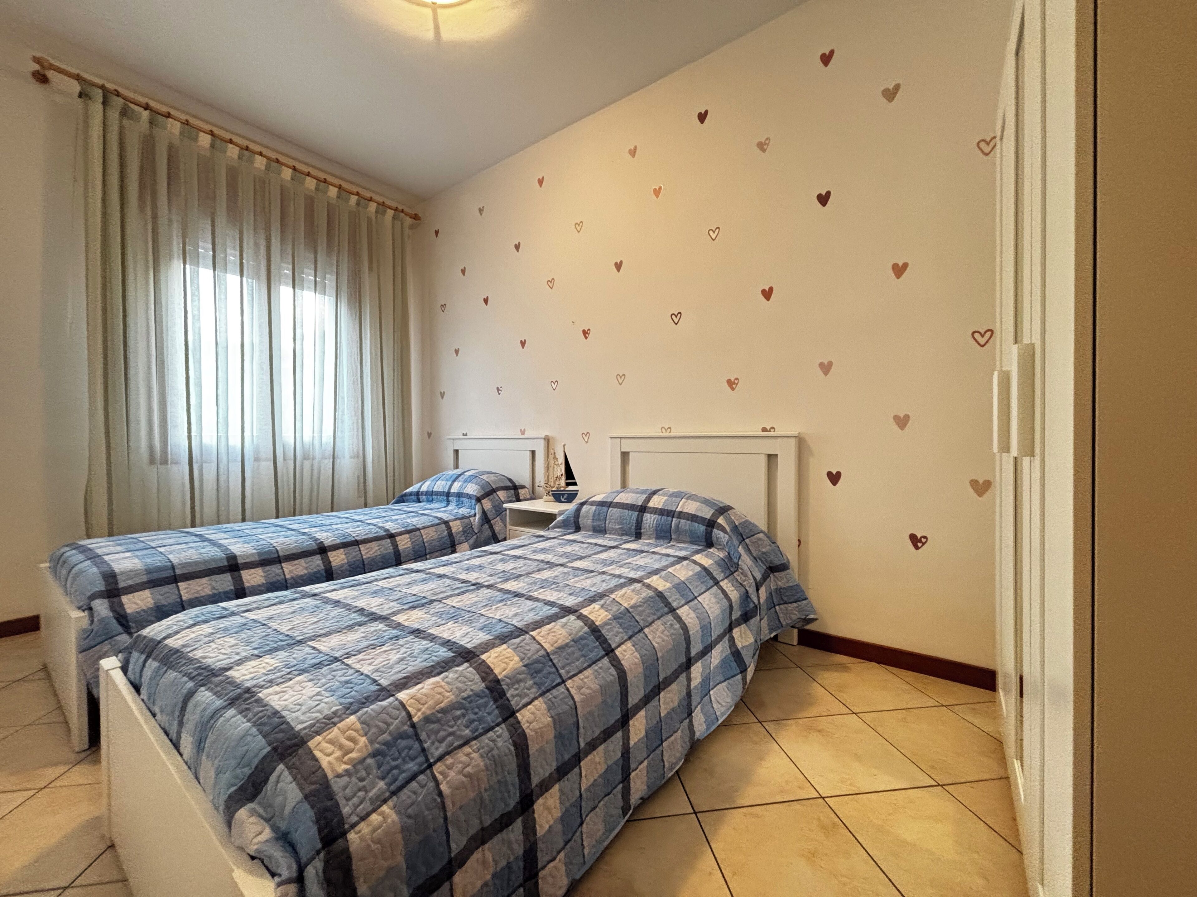 Een slaapkamer met twee bedden, een raam met gordijnen, een nachtkastje met een lamp en een muur versierd met hartjes.