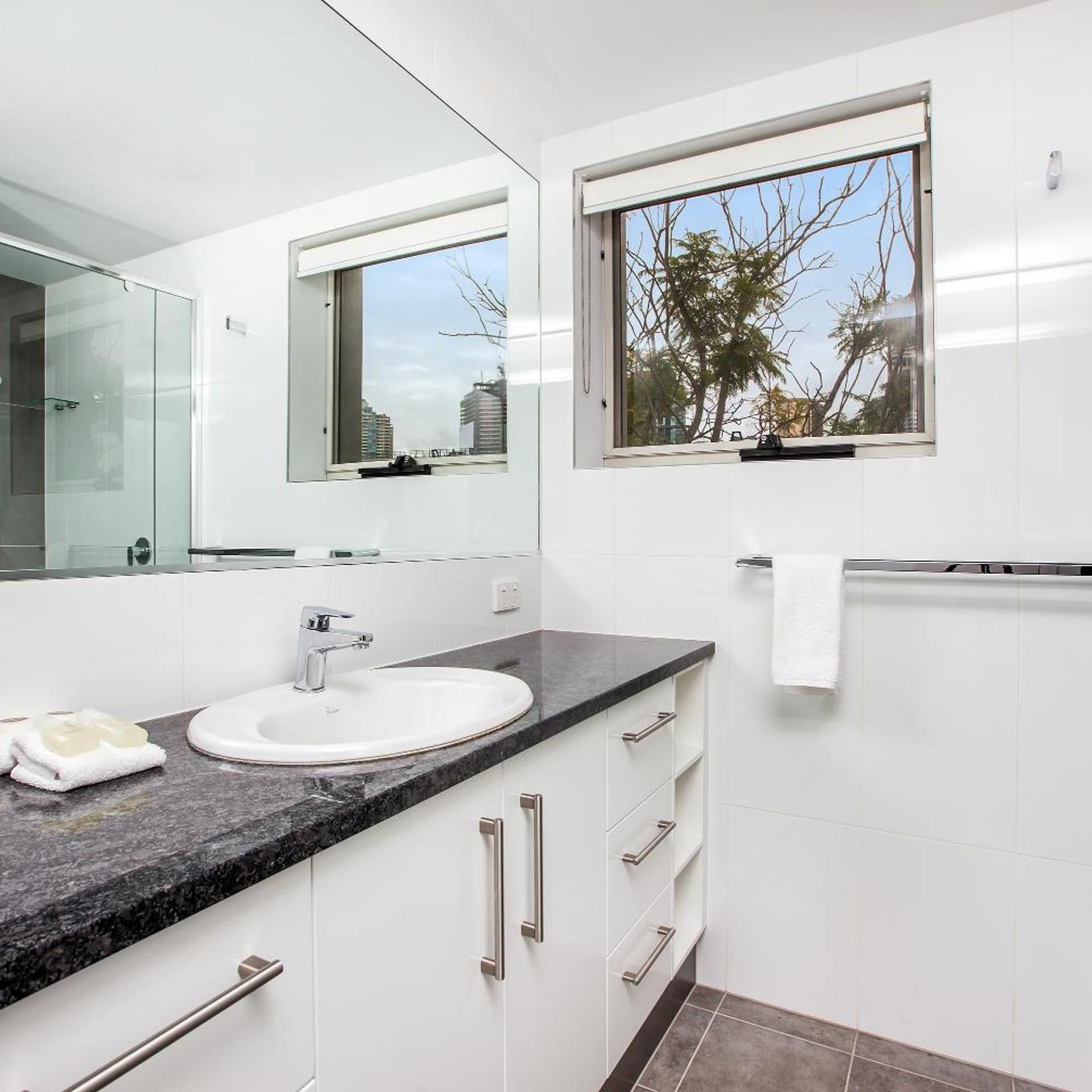A modern bathroom with a white sink, a large mirror, and a window with a view of trees and buildings.