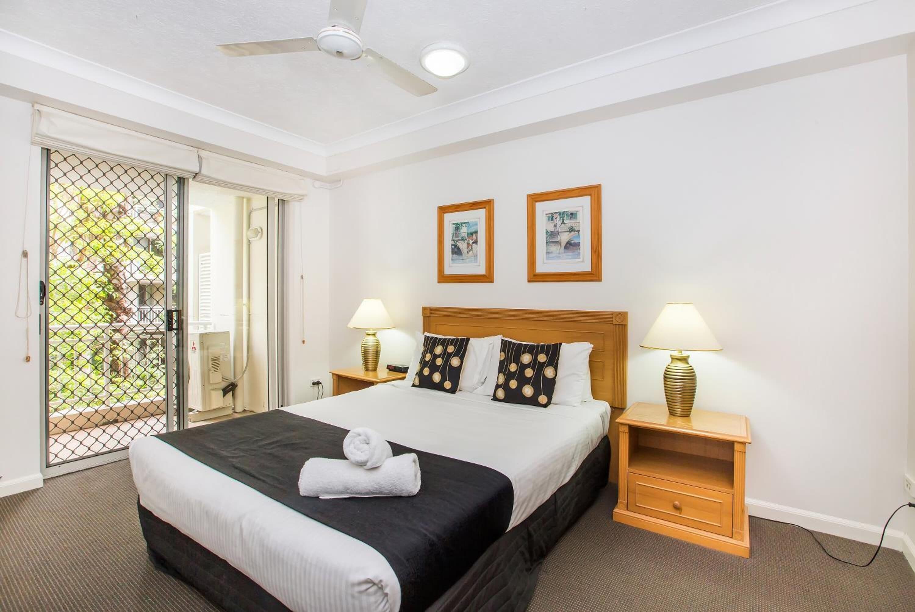 Quarto de hotel com uma cama grande, duas mesas de cabeceira com luminárias, um ventilador de teto e uma porta de vidro deslizante para o exterior.