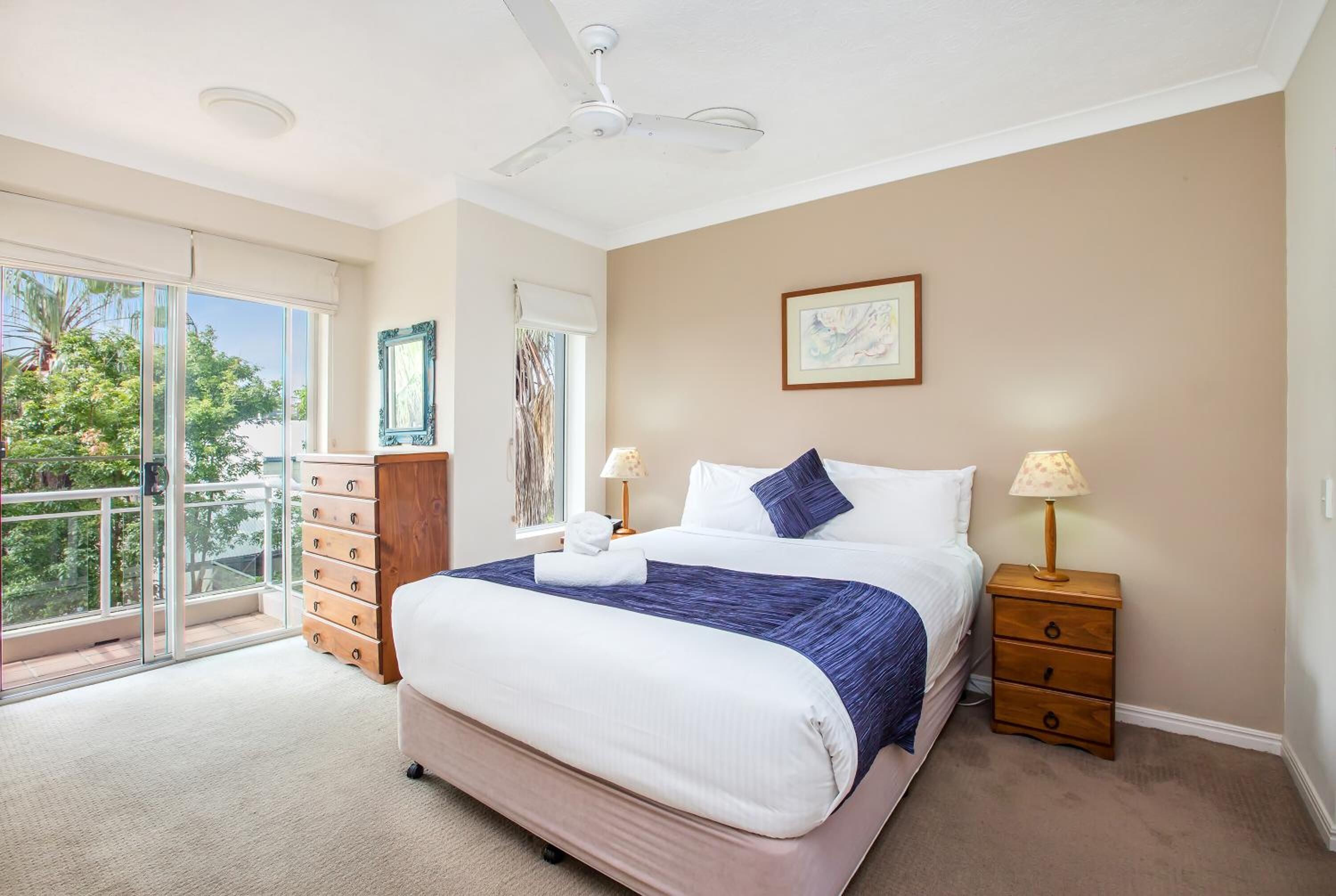 A bedroom with a large bed, a ceiling fan, a nightstand with a lamp, and a view of the outdoors through a sliding door.