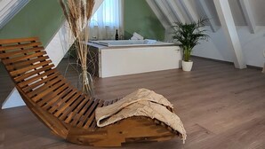 Un fauteuil en bois dans une chambre à coucher aménagée en loft, avec un plafond en pente, une petite table de chevet, une plante en pot et un vase contenant des plantes séchées.