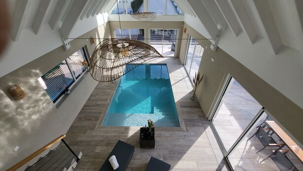 Une vaste piscine intérieure, offrant une vue dégagée sur le ciel, est entourée d’une architecture moderne avec de grandes fenêtres et un plancher en bois.