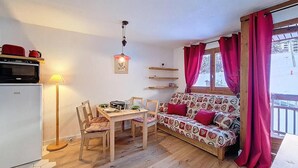 A cozy room with a wooden floor, a dining table with chairs, a sofa with patterned cushions, a small fridge, a lamp, and a window with curtains.