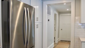 Une cuisine avec des meubles blancs, un réfrigérateur en acier inoxydable, un plancher en bois et une porte blanche donnant sur une autre pièce.