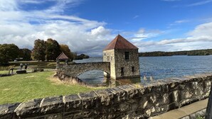 A stone structure with a red roof by a body of water, surrounded by a stone wall and grassy area. - Quaint Cabin #7 - Pymatuning Lake (Jamestown)