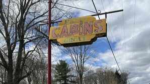 A yellow sign with "Maple Cabins Grove" written on it, a red pole, and a grassy area with white flowers. - Quaint Cabin #7 - Pymatuning Lake (Jamestown)