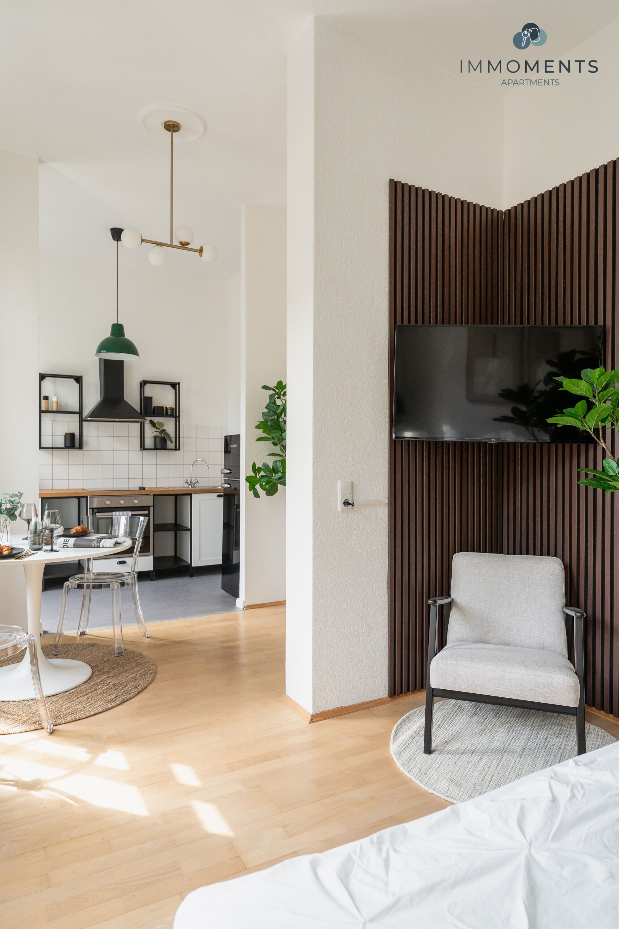 Ein modernes Wohnzimmer mit Sofa, Essbereich mit Tisch und Stühlen, Küche und einem an der Wand befestigten Fernseher, sowie einer Pflanze.