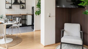 Een moderne woonkamer met een bank, een eethoek met tafel en stoelen, een keuken met een aan de muur bevestigde televisie, en een plant.
