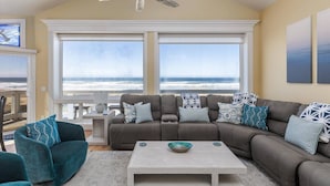 Ein Wohnzimmer mit einem großen Fenster mit Blick auf den Strand, einem Deckenventilator und einem Ecksofa mit gemusterten Kissen.