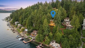 Une vue aérienne d’une propriété côtière comprenant plusieurs maisons entourées d’une forêt dense, reliées par des passerelles en bois, et des bateaux amarrés le long du rivage.