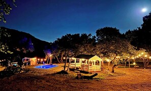 A small, illuminated gazebo-like structure with a pool, surrounded by trees and a mountain in the background.