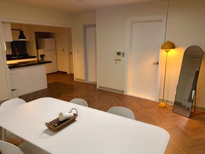 Eine kompakte Küche mit Kühlschrank, Mikrowelle und Kaffeemaschine. Ein Esstisch mit Stühlen und ein Korb mit einer Tasse stehen bereit. An der Wand befinden sich ein Spiegel und eine Lampe.