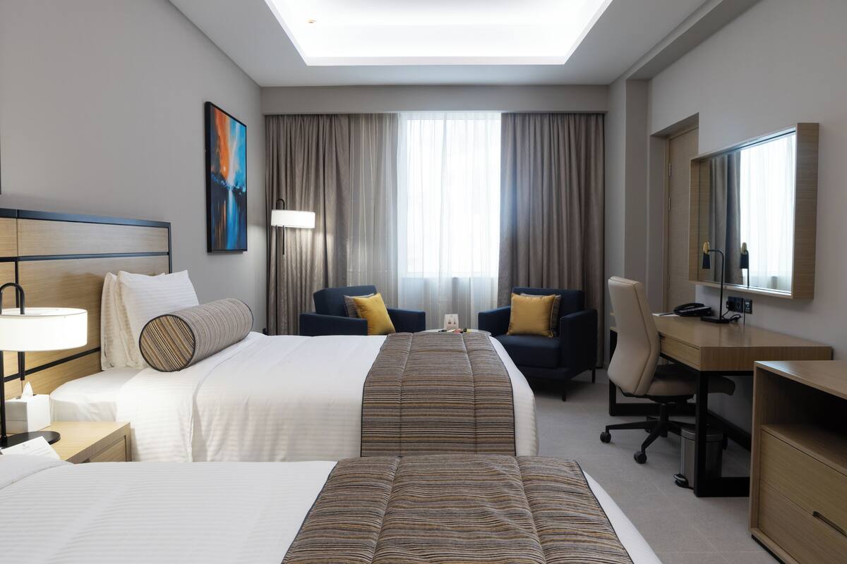 Une chambre d’hôtel avec deux lits, un bureau, une chaise, une lampe et une grande fenêtre avec des rideaux.