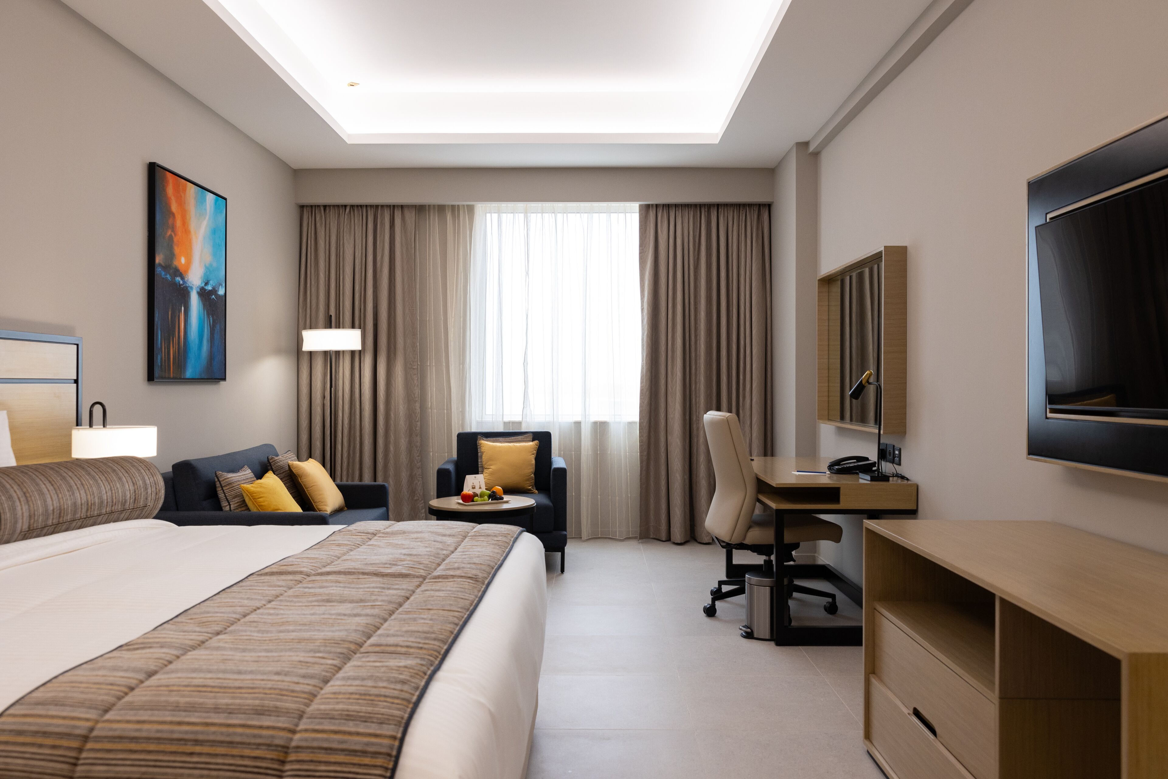 A hotel room with a large bed, a desk, a chair, a TV, and a window with curtains.