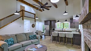 A spacious living area with a high ceiling, wooden beams, a staircase, a kitchen with bar stools, and a dining area.