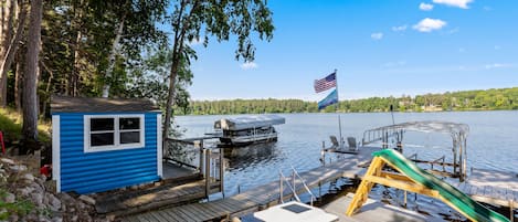 A dock with a small blue shed, a green slide, and a boat on the water.