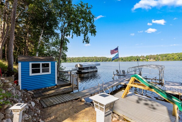 A dock with a small blue shed, a green slide, and a boat on the water.